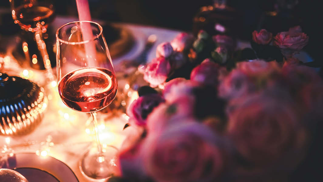 L'immagine mostra un bicchiere di vino rosato su un tavolo decorato con fiori rosa e luci calde, creando un'atmosfera elegante e intima. L'immagine mostra un bicchiere di vino rosato su un tavolo decorato con fiori rosa e luci calde, creando un'atmosfera elegante e intima.