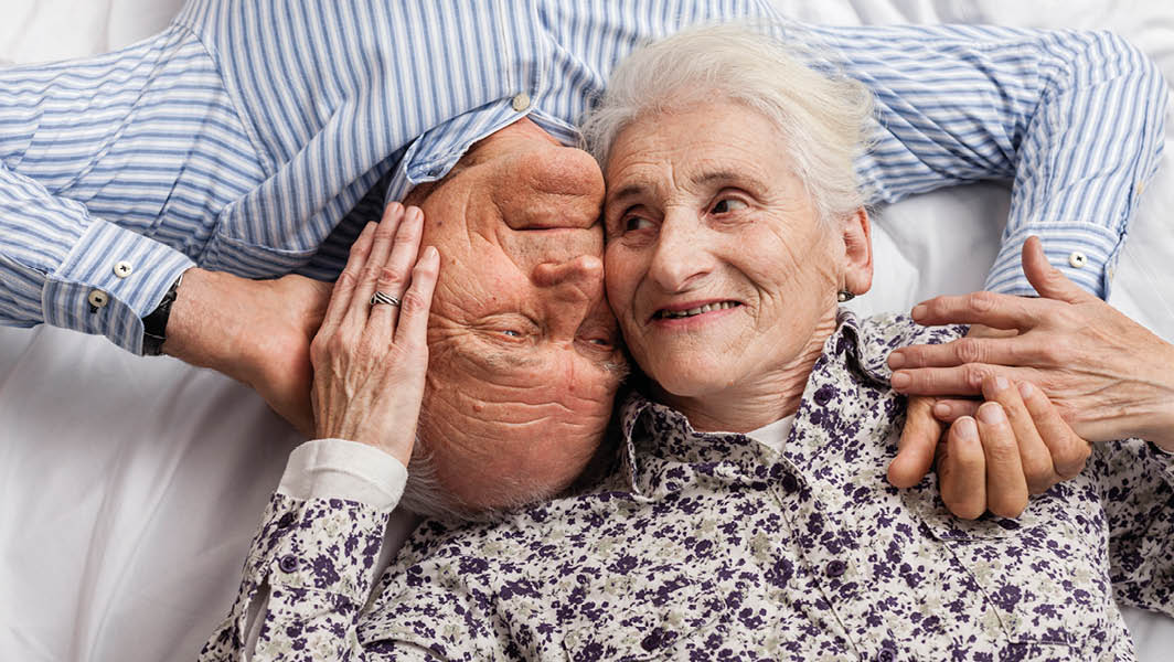 Una coppia di anziani distesi uno accanto all'altra, sorridenti e sereni, raffiguranti il concetto di benessere senior. Entrambi indossano abiti comodi, l'uomo con una camicia a righe e la donna con una camicia a fiori.