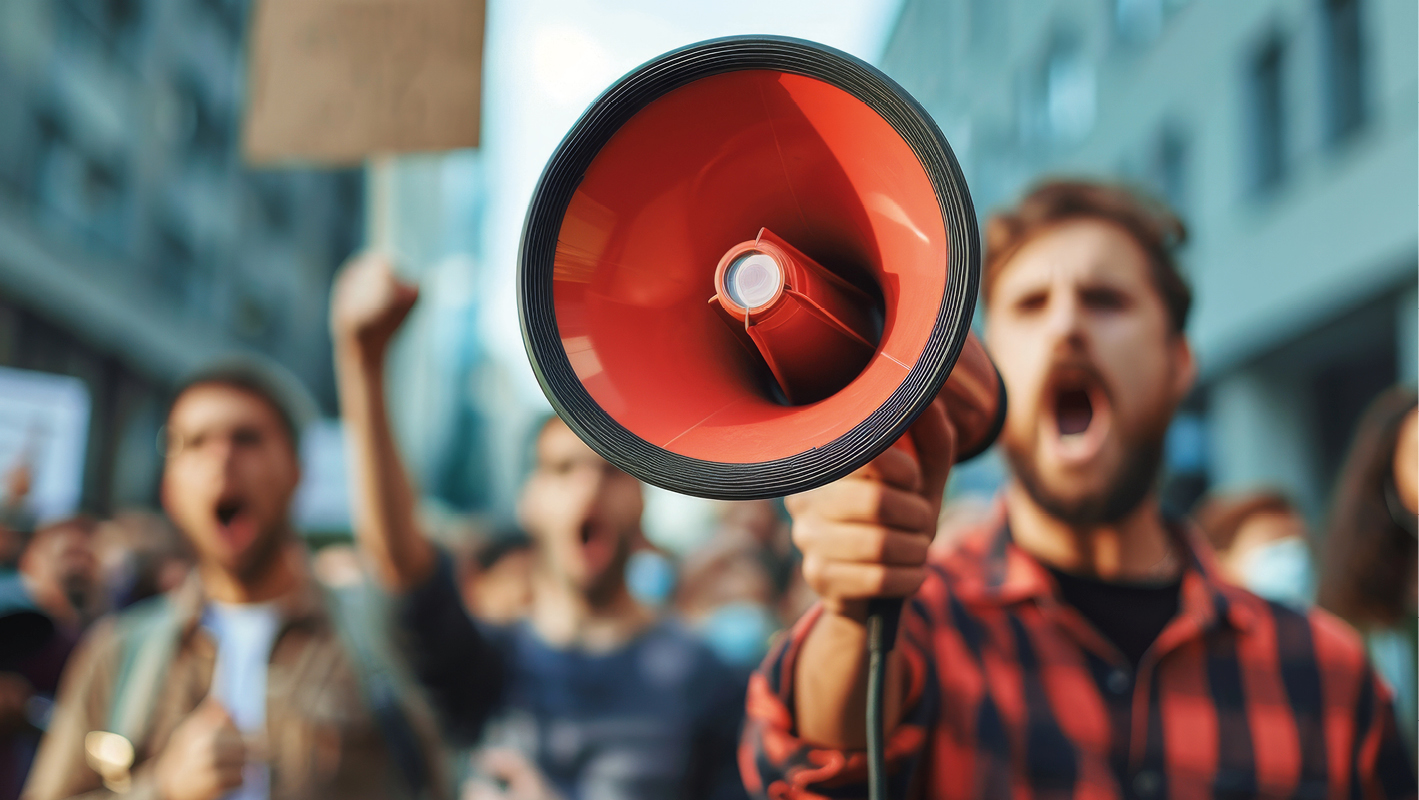 Un uomo tiene in mano un megafono rosso durante una manifestazione di protesta contro l'inceneritore, mentre urla insieme ad altre persone sfocate sullo sfondo.