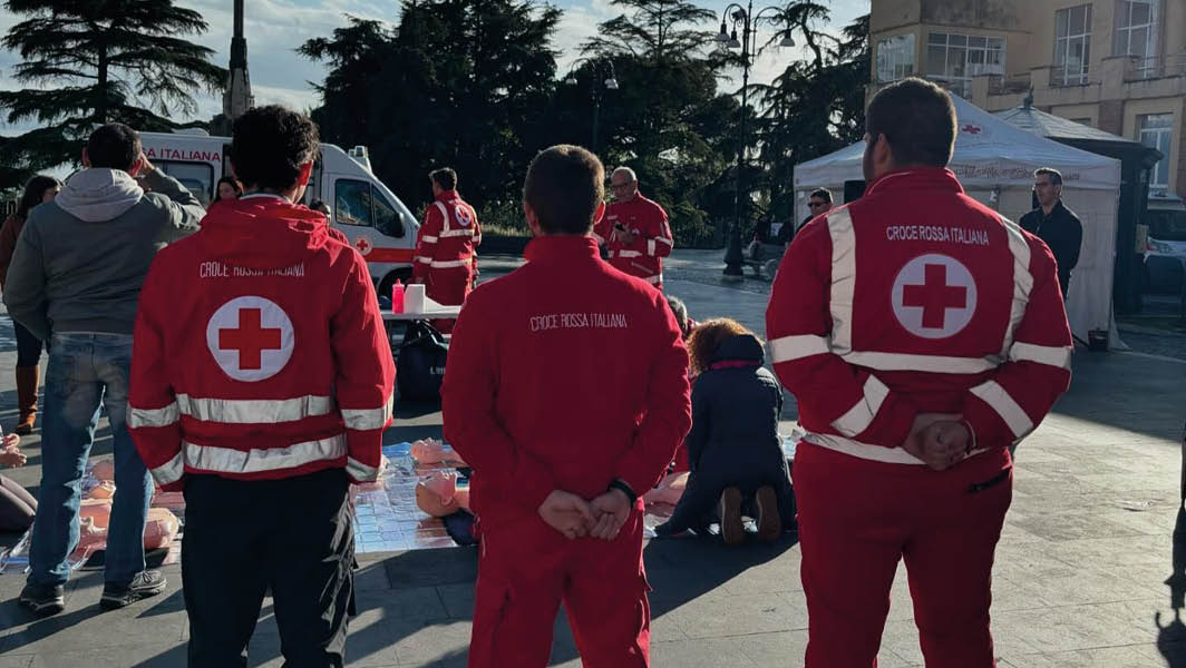 Volontari della Croce Rossa Italiana durante una dimostrazione pubblica all'aperto, con manichini per addestramento e un'ambulanza sullo sfondo.