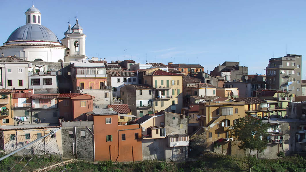 Veduta del centro storico di Ariccia con case colorate su una collina e la cupola della Chiesa di Santa Maria Assunta sullo sfondo, sotto un cielo limpido.