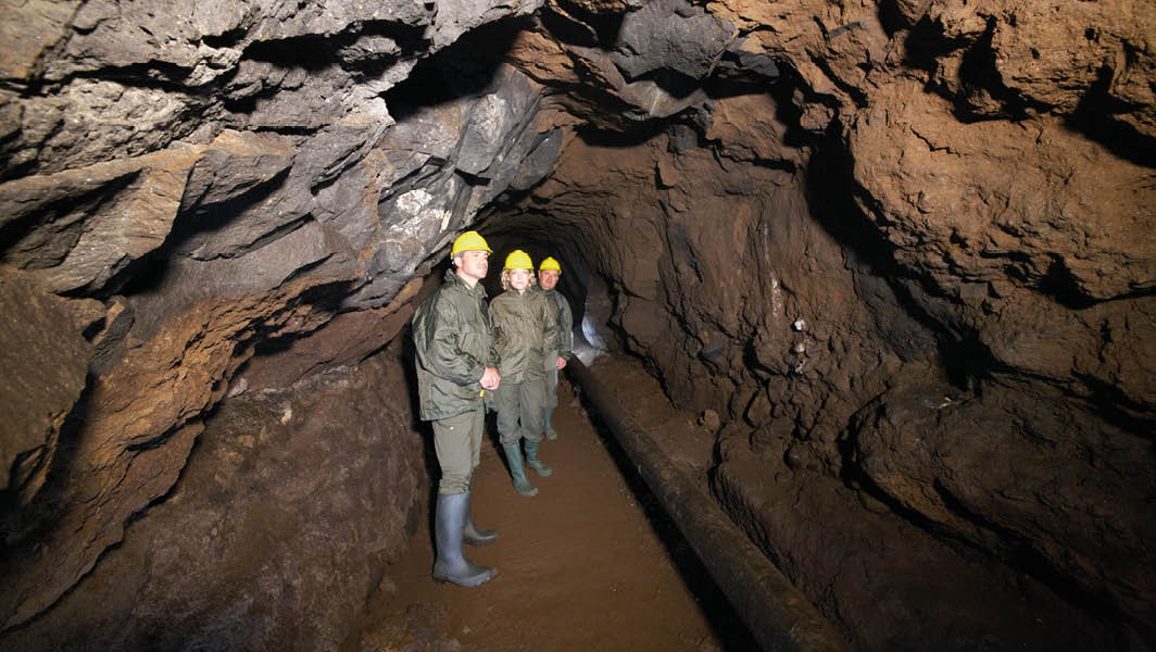 Esploratori all'interno dell'antico emissario del lago di Nemi, indossano stivali e caschi di sicurezza in un ambiente sotterraneo roccioso.