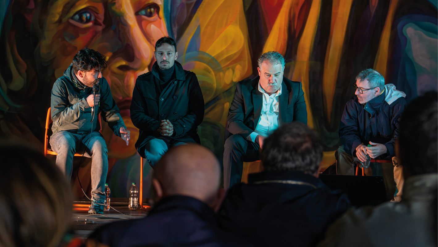 Quattro uomini seduti su un palco durante un dibattito, con un murale colorato che raffigura un volto umano alle loro spalle, tra cui Edoardo Menicocci, segretario del PD Velletri, e Orlando Pocci, ex sindaco di Velletri.