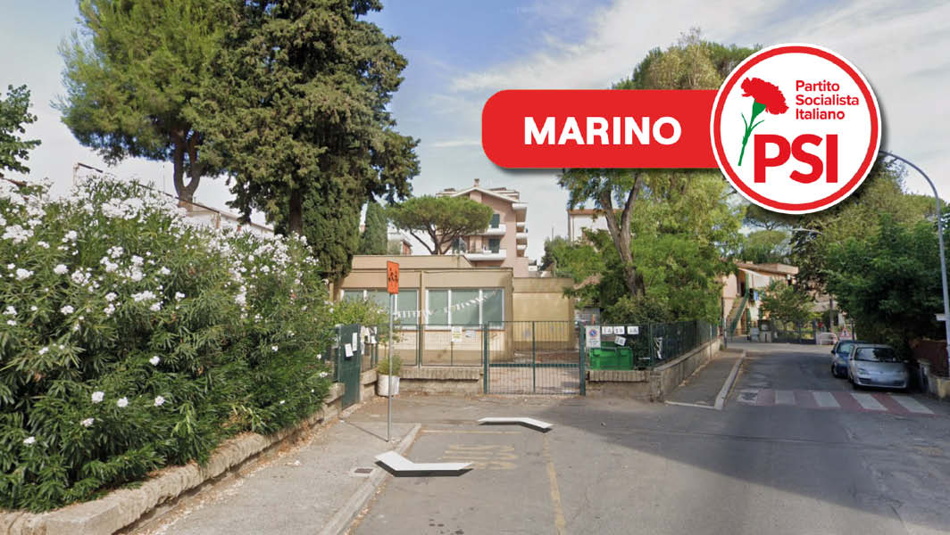 Ingresso della scuola dell'infanzia Elsa Morante a Frattocchie, Marino, con un cancello chiuso, piante rigogliose a lato e un edificio residenziale sullo sfondo.