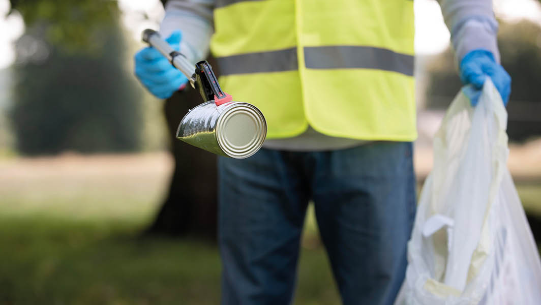 Operatore ecologico che raccoglie una lattina a Monte Porzio Catone con un attrezzo di presa, indossando un giubbotto ad alta visibilità e guanti, con un sacchetto per rifiuti in mano.