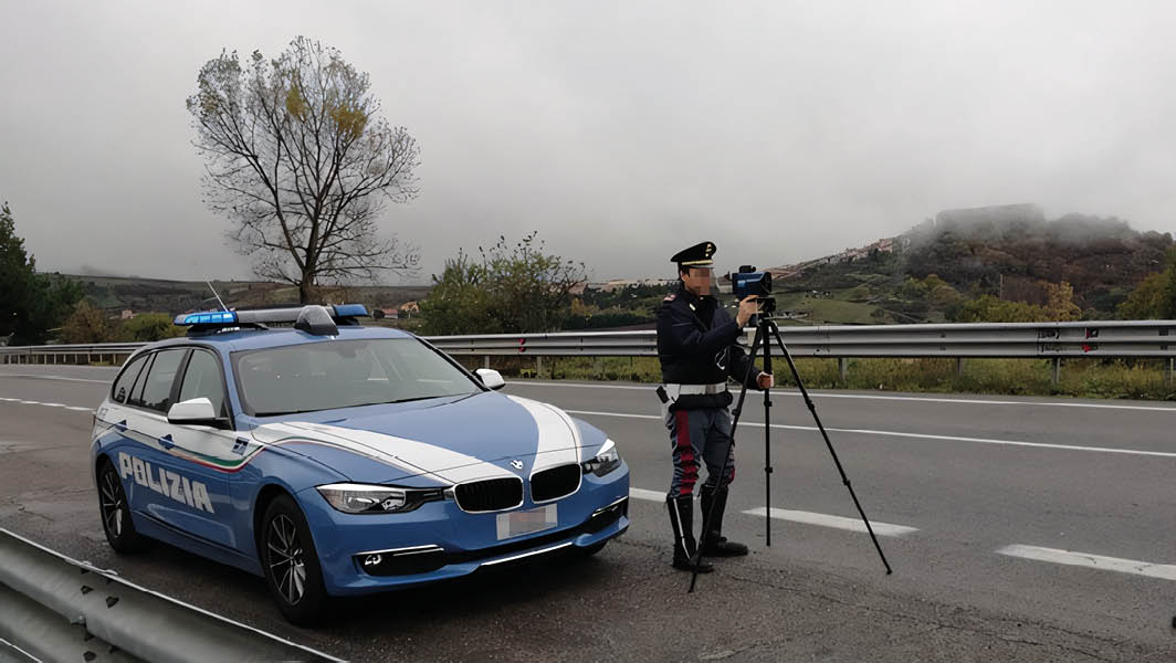 Agente della polizia stradale italiana con autovelox portatile accanto a un'auto di pattuglia su una strada.