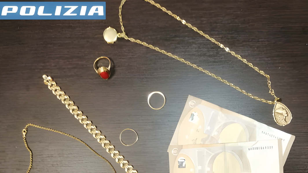 Un'immagine che mostra gioielli e denaro recuperati dalla Polizia di Stato in un'operazione contro truffe agli anziani. Tra gli oggetti recuperati vi sono una collana, un anello con pietra rossa, una fede, altri anelli d'oro e alcuni biglietti. Nella parte superiore dell'immagine è visibile il logo della Polizia.