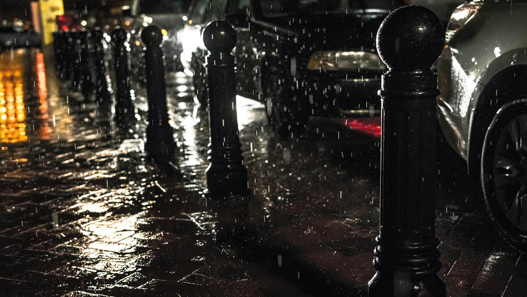 Marciapiede bagnato dalla pioggia, con paletti neri e auto parcheggiate lungo la strada di notte, una situazione simili a quelle che si potrebbero verificare a Marino.