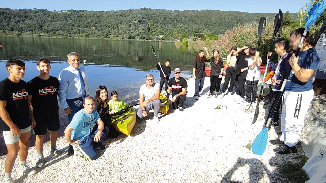 Gruppo di studenti e istruttori del Centro Canoe di Nemi, insieme a rappresentanti locali, posano sulla riva del lago di Nemi con canoe e attrezzature sportive.