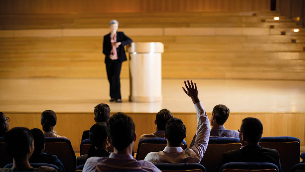 Un incontro pubblico con una relatrice sul palco e partecipanti seduti in una sala conferenze, uno dei quali alza la mano per intervenire.