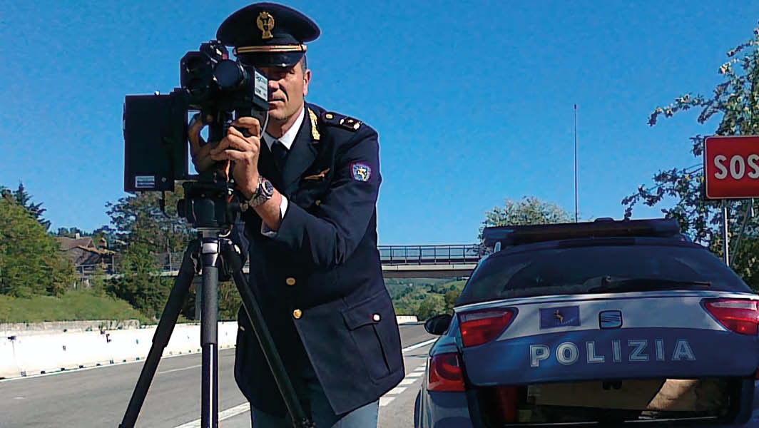 Agente della Polizia di Stato con autovelox su treppiede in una strada del Lazio, accanto a una pattuglia