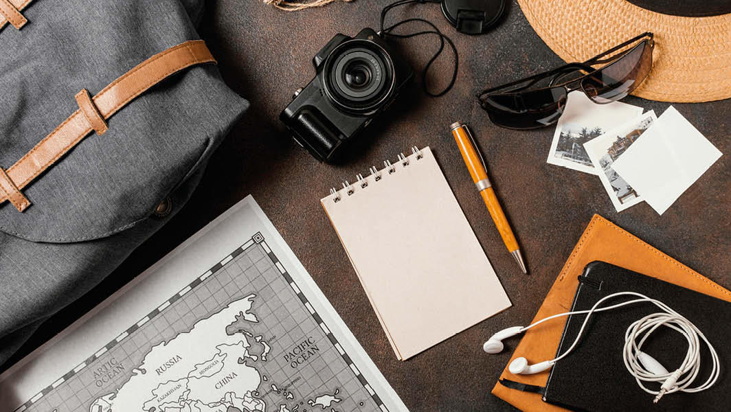 Oggetti da viaggio disposti su una superficie: zaino, mappa, fotocamera, cappello, occhiali da sole, bloc-notes e penna, simboli dell'organizzazione di un viaggio. Riferimento al Civitavecchia Shuttle Express.