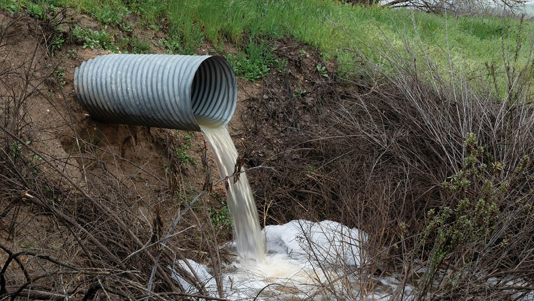 Un tubo di scarico rilascia acque reflue nel terreno, similarmente a quanto accade a Frattocchie, evidenziando i problemi ambientali della zona.