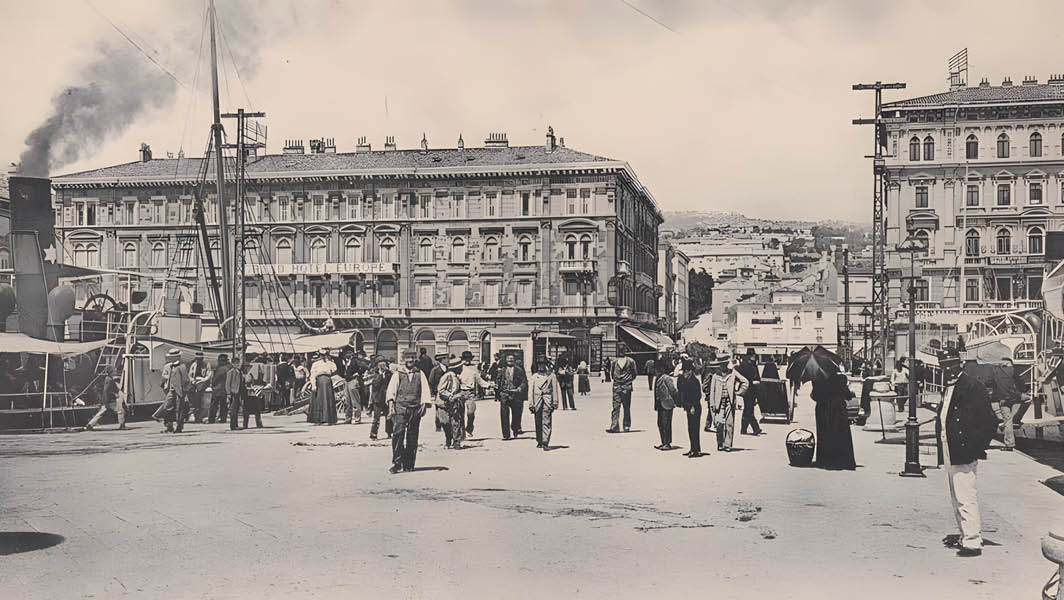 Fotografia d'epoca di una piazza di Fiume con persone in abiti storici, edifici imponenti e un porto con navi a vapore sullo sfondo.