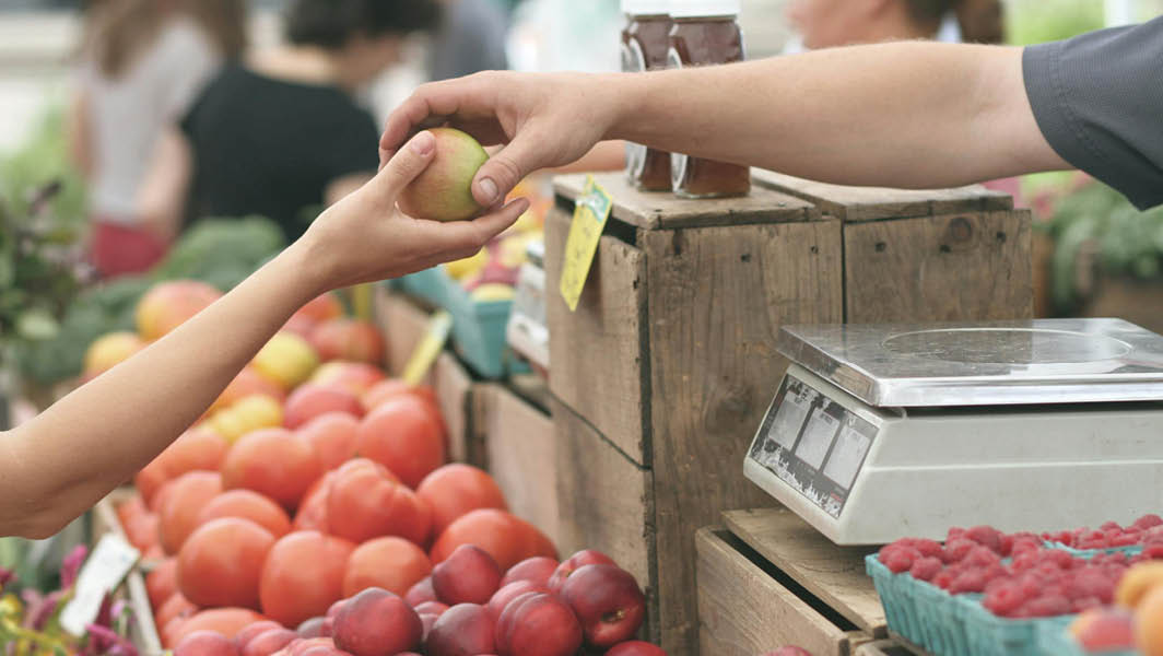 Scambio di una mela tra venditore e cliente in un mercato all'aperto come quello di Rocca di Papa, con cassette di frutta fresca esposta.