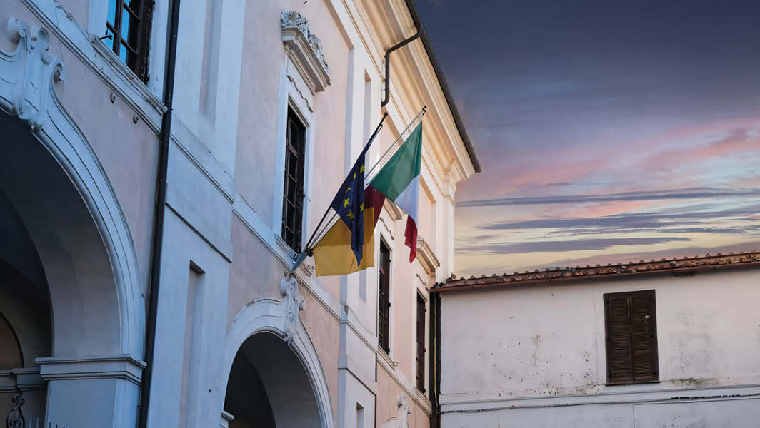 Facciata di Palazzo Savelli ad Albano Laziale con bandiere italiane, europee e comunali al tramonto.