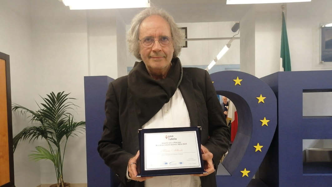 Mario Alberti premiato con la Targa Eccellenze Europee, tiene in mano il riconoscimento durante la cerimonia presso la sede del Parlamento Europeo a Roma, con il logo dell'evento sullo sfondo.