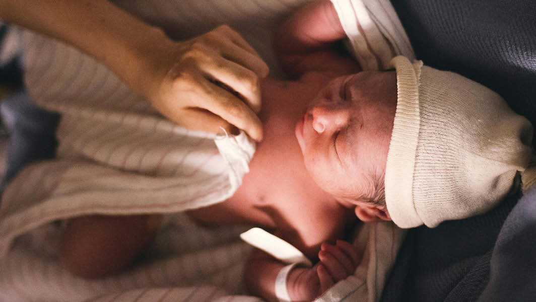 Un neonato avvolto in una coperta, con un cappellino bianco, accudito da una mano gentile. L'immagine simboleggia la riapertura del punto nascita presso l'ospedale Paolo Colombo di Velletri.