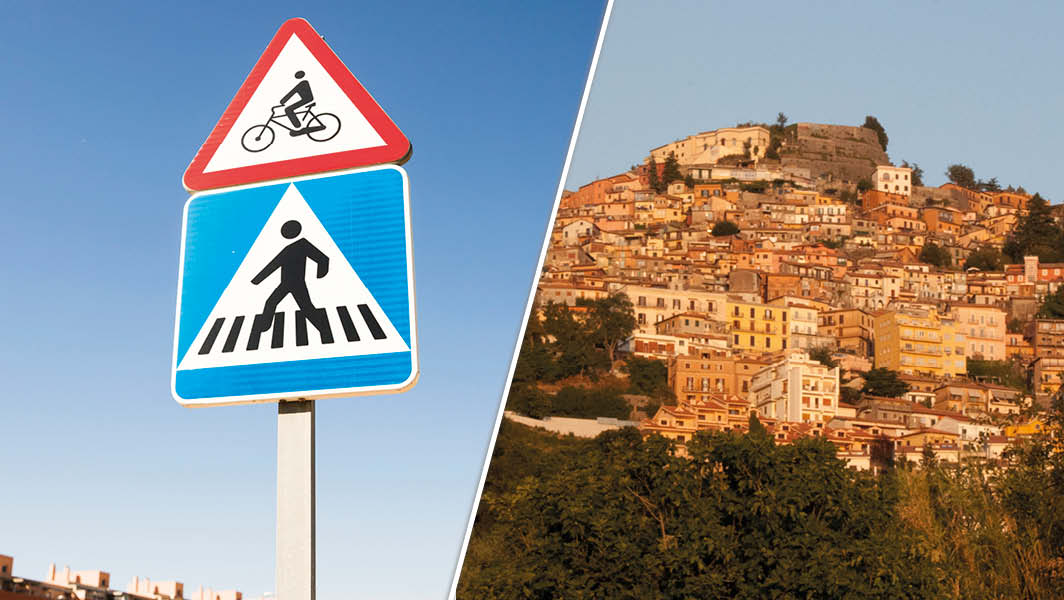 Cartello stradale con segnaletica per attraversamento pedonale e ciclisti a Rocca di Papa, con vista panoramica del borgo storico arroccato sul monte.