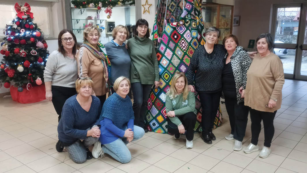 Un gruppo di donne di Rocca di Papa posa davanti a un albero di Natale decorato con quadrati di lana colorati, simbolo di una tradizione collettiva e di solidarietà.