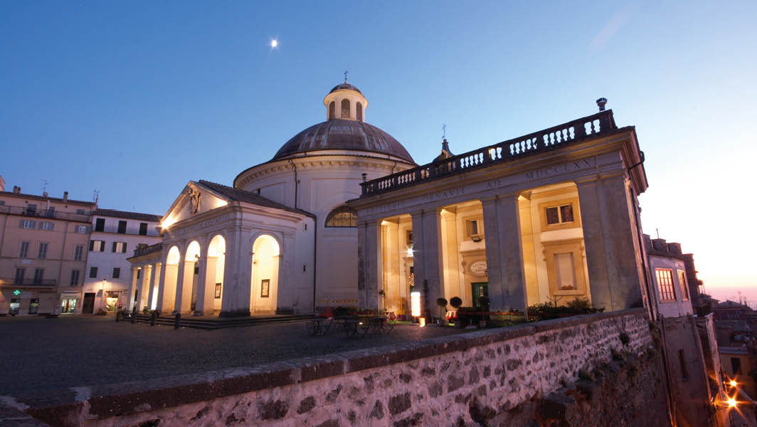 La Collegiata di Santa Maria Assunta ad Ariccia illuminata di sera, con la piazza antistante e il cielo al crepuscolo.