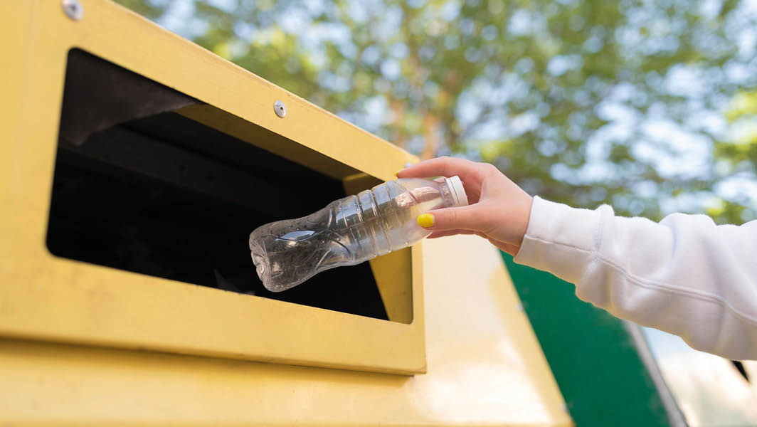 Mano che inserisce una bottiglia di plastica in un contenitore giallo per la raccolta differenziata, con sfondo sfocato di alberi.