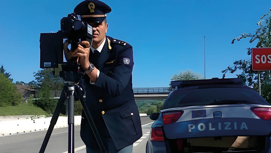 Agente della Polizia di Stato in uniforme mentre utilizza un autovelox mobile su una strada, con un'auto della polizia sullo sfondo e un cartello SOS visibile.