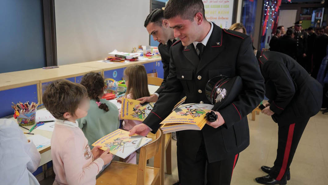 Un giovane Carabiniere consegna un libretto illustrato a un bambino durante una visita ai reparti pediatrici, con altri Carabinieri impegnati nello stesso gesto di solidarietà.