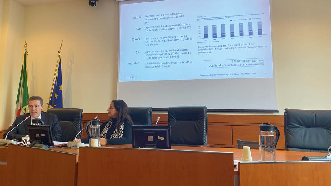 Sala conferenze con due relatori seduti, bandiere istituzionali sullo sfondo e una presentazione proiettata sul tema della crisi idrica; il posto riservato al Comune di Genzano di Roma è vuoto.