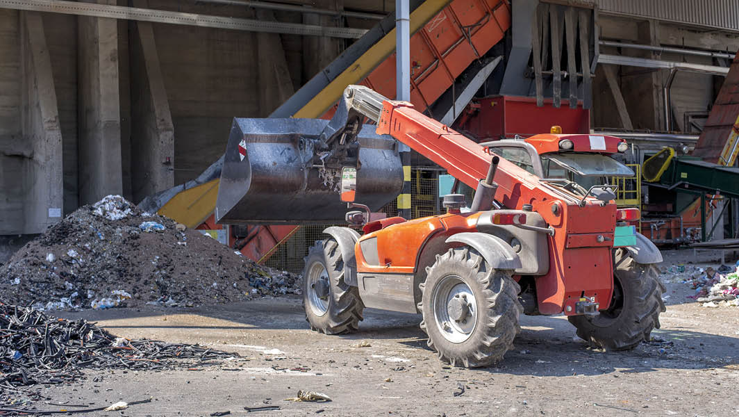 Macchinario edile in un cantiere impegnato nel sollevamento di materiali di scarto, con cumuli di rifiuti edili sullo sfondo.