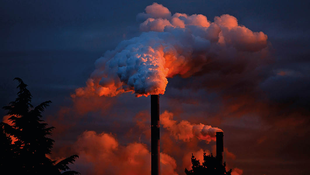 Due ciminiere di un inceneritore emettono fumo denso e illuminato dal tramonto, mentre il cielo scuro incombe sullo sfondo.