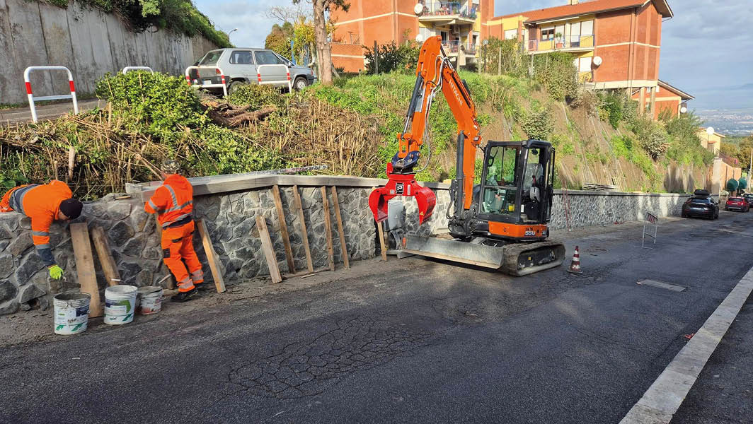 Operai al lavoro su un muro in pietra a Monte Porzio Catone con un escavatore in azione sulla strada.