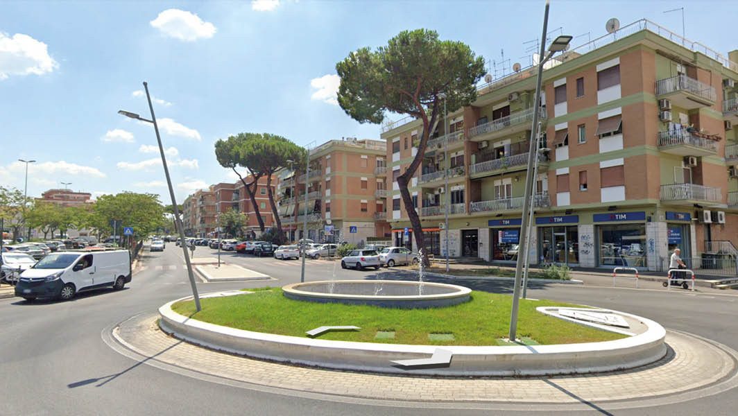 Rotatoria con fontana e pini a Viale Kennedy, Ciampino, con edifici residenziali sullo sfondo.