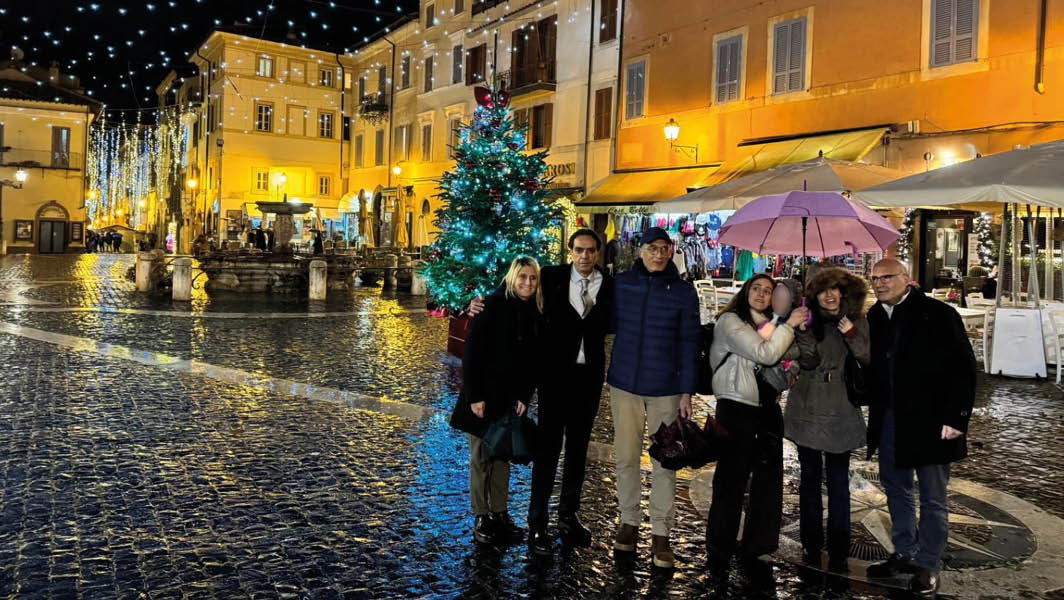 Persone riunite nella Piazza della Libertà a Castel Gandolfo, con l’albero di Natale illuminato e le luci festive che decorano il borgo.