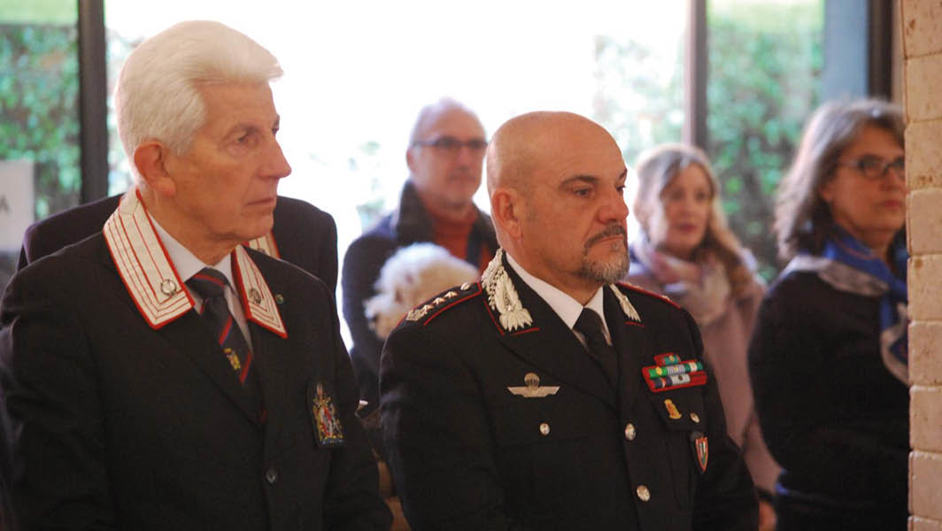 Due ufficiali dei Carabinieri partecipano alla cerimonia del Precetto natalizio dell'Associazione Nazionale Corazzieri. Sullo sfondo, altre persone assistono con attenzione.