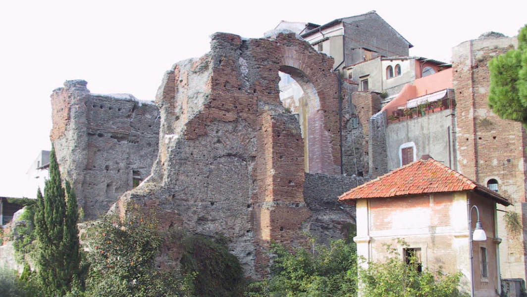 Rovine delle Terme di Caracalla ad Albano Laziale, con strutture in mattoni antichi e resti archeologici circondati da vegetazione e edifici storici.